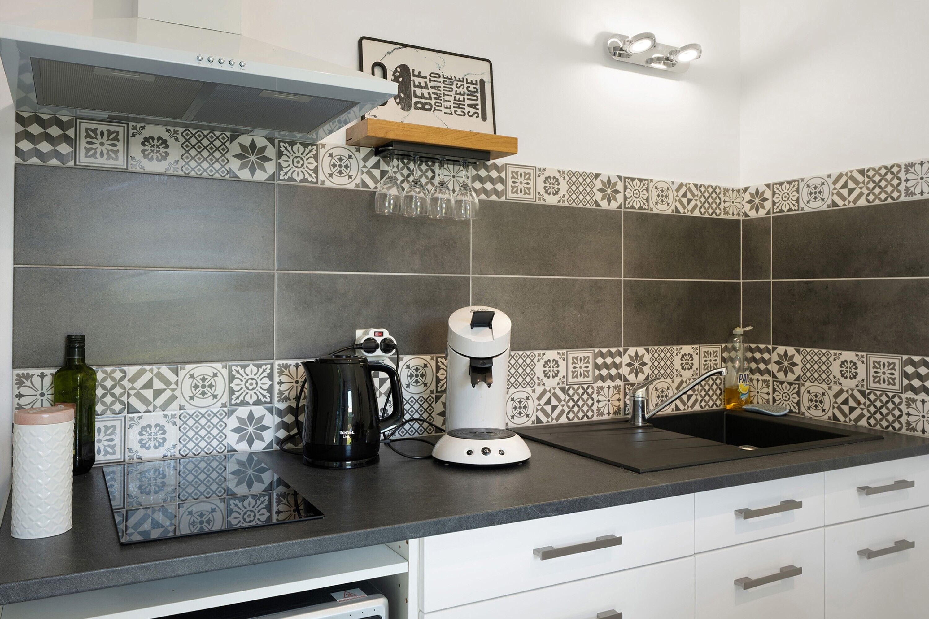 Une cuisine avec des carreaux décoratifs, une machine à café, une bouilloire et une bouteille.