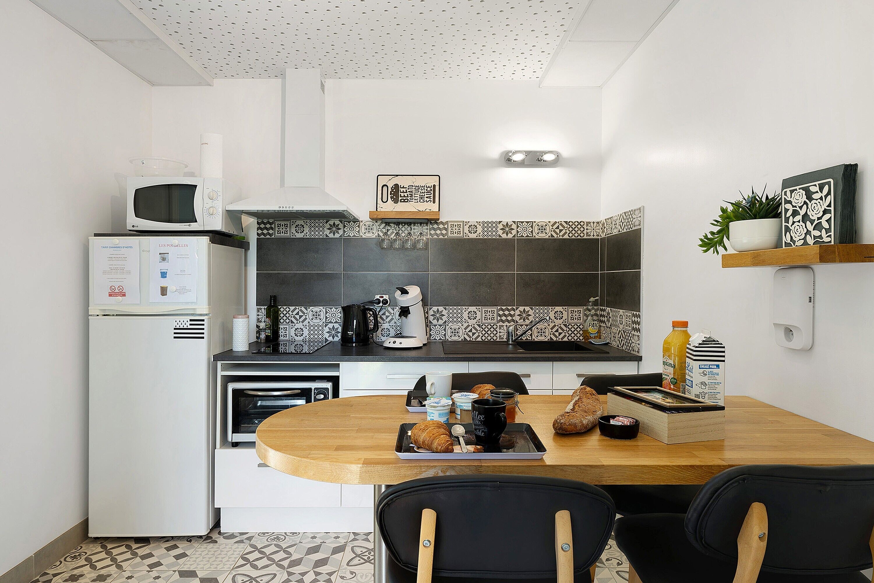 A compact kitchen with a microwave, coffee maker, and toaster, a dining table with chairs, and a decorative shelf with a plant.