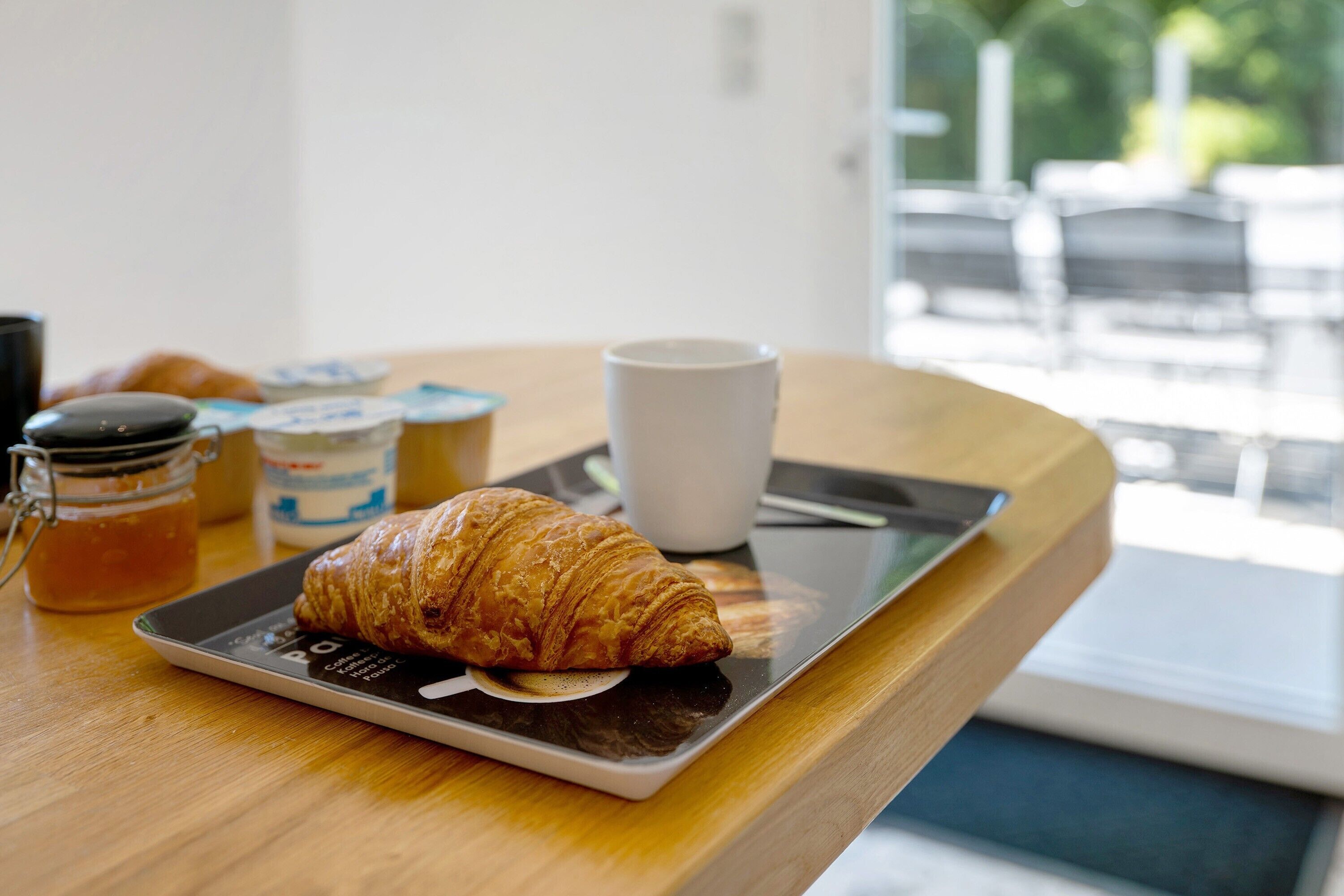 A croissant on a tablet, a cup, and jars of honey and jam on a wooden table.