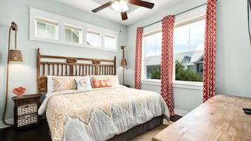 Un dormitorio con cabecera de madera, cama con ropa de cama estampada, una mesa de madera, ventilador de techo y vistas a través de una ventana con cortinas rojas.