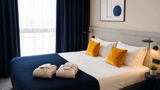 A hotel room with a large bed, a headboard, a bedside lamp, a potted plant, and a window with curtains. - Cobalt36 (Bolesławiec)