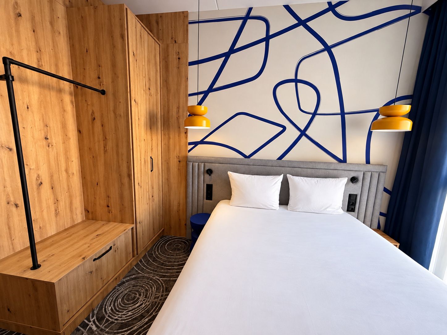 Une chambre d’hôtel avec un lit, une armoire en bois et un mur orné de lignes bleues abstraites.