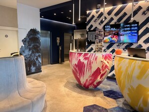 Una recepción moderna con una gran escultura abstracta, un reloj digital que muestra la disponibilidad de habitaciones y un área de descanso con un sofá.