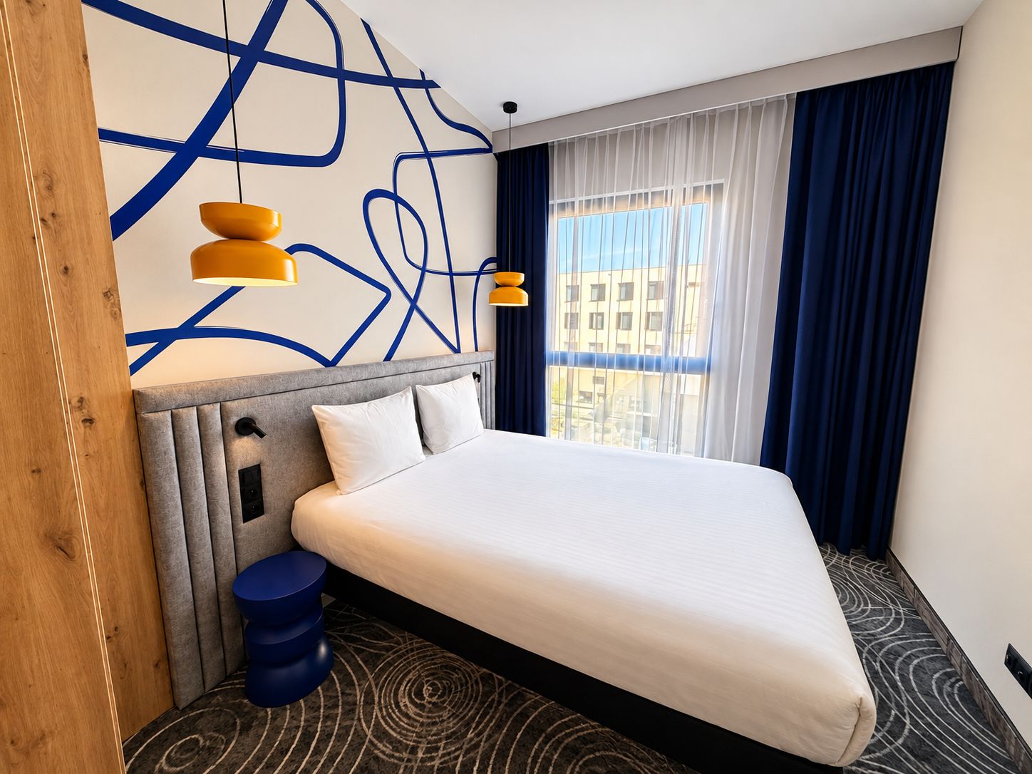 Habitación de hotel con una cama grande, cabecera con panel de control integrado, pared azul con líneas abstractas, ventana con cortinas y alfombra estampada.