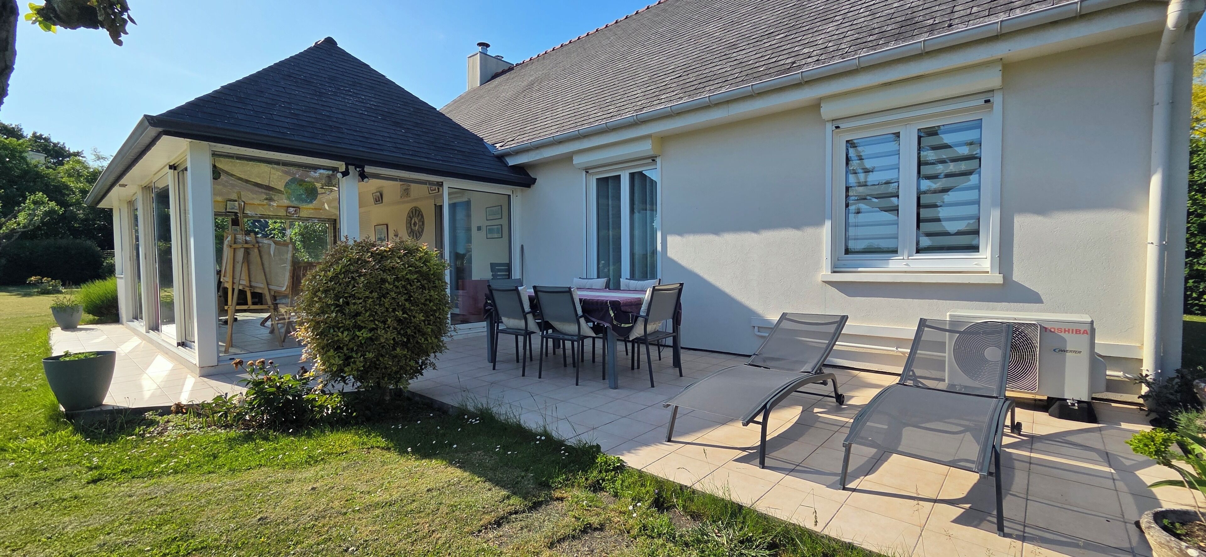 Ein Haus mit überdachter Terrasse, einem Essbereich mit Stühlen, einem kleinen Garten und einer Klimaanlage.