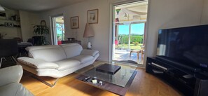 A living room with a white sofa, a glass coffee table, a TV, and a large window with a view of the outdoors. - Beautiful, Spacious Single-Story House with a Large Enclosed Garden in Trégunc (Trégunc)