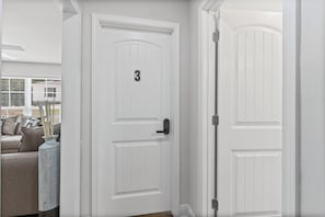 Una puerta blanca con el número 3 y una cerradura electrónica, otra puerta blanca con un pestillo, y un sofá beis con cojines.