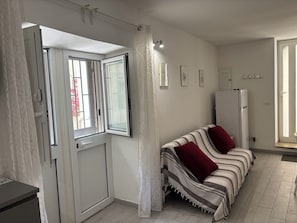 A room with a sofa, a refrigerator, a door with a window, and a wall with framed pictures. - DIONE: A cozy, fully equipped apartment in the heart of Ortigia, Syracuse (Siracusa)