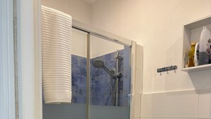 A bathroom with a glass-enclosed shower, a white sink, and a shelf with toiletries. - DIONE: A cozy, fully equipped apartment in the heart of Ortigia, Syracuse (Siracusa)
