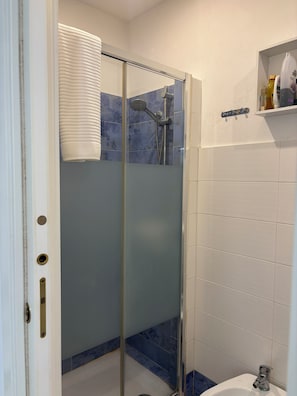 A bathroom with a glass-enclosed shower, a white sink, and a shelf with toiletries. - DIONE: A cozy, fully equipped apartment in the heart of Ortigia, Syracuse (Siracusa)