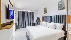 Ein modernes Hotelzimmer mit einem großen Bett, einem flachbildfernseher an der Wand, einem Schreibtisch mit Stuhl und einem fenster mit Vorhängen.
