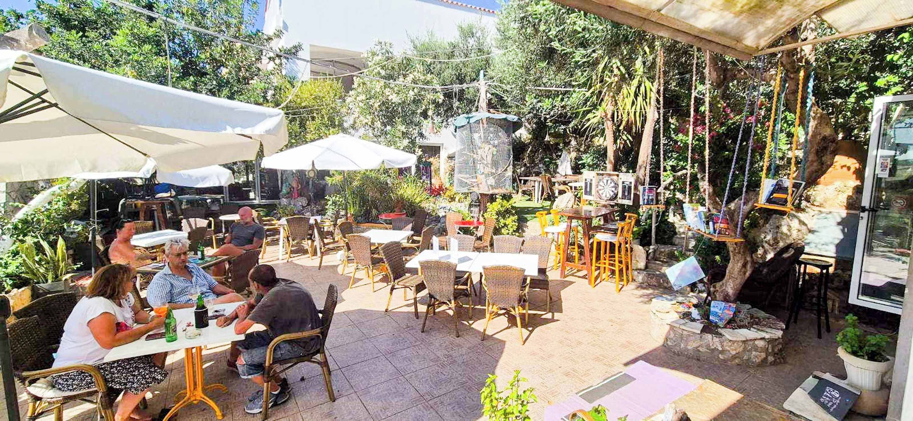 Un cafè a l'aire lliure amb taules i cadires, envoltat de vegetació i una formació rocosa.