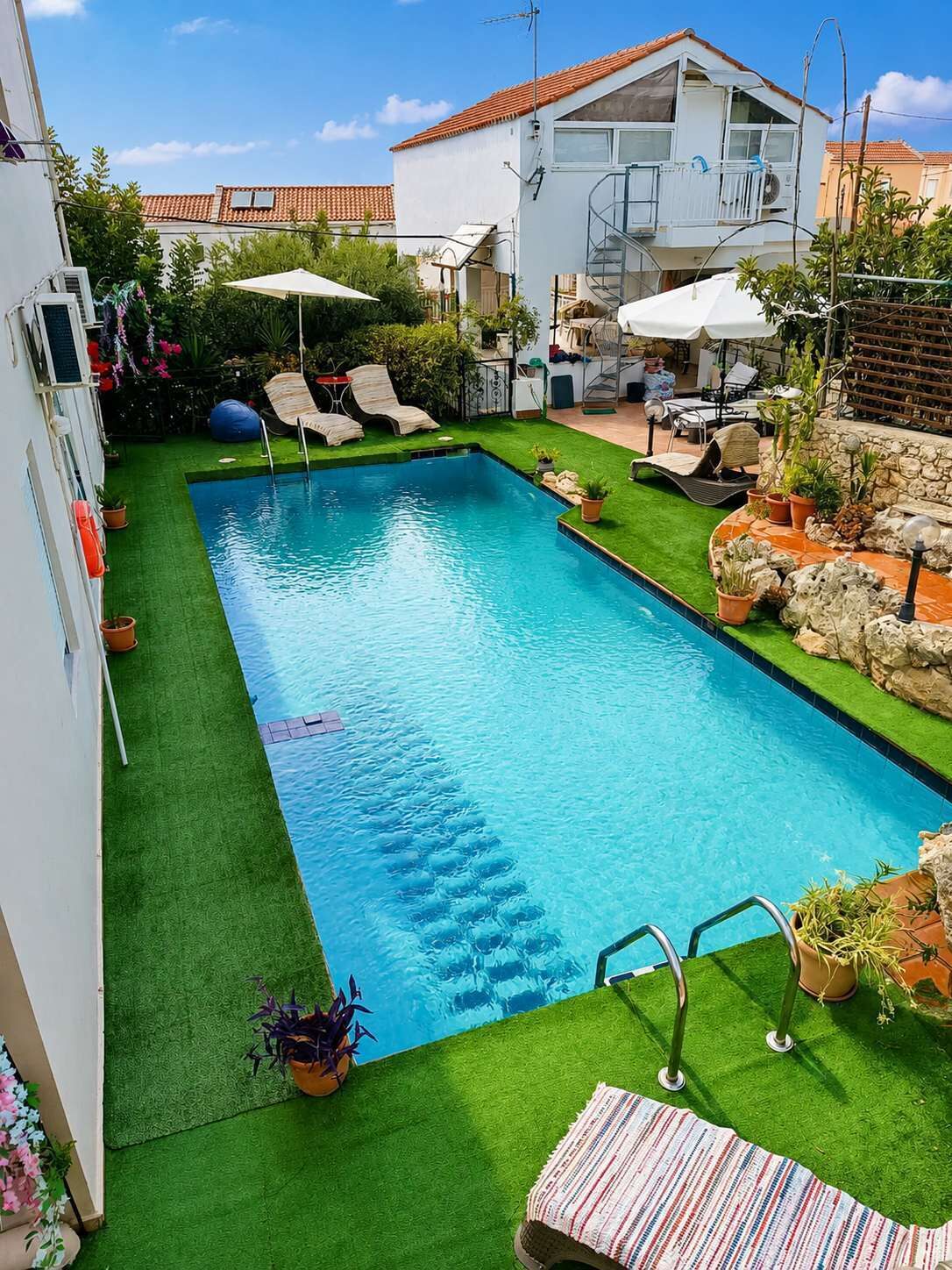 Ein rechteckiger Pool mit glasklar blauem Wasser, umgeben von künstlichem grünen Rasen, neben einem weißen Gebäude mit einem Ziegeldach.