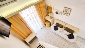 A hotel room with a sofa, a small table, a bedside table, a TV, a window with curtains, and a wall-mounted air conditioning unit. - Bicorna Chania Pool Escape - Yellow Room 1 (Plaka)