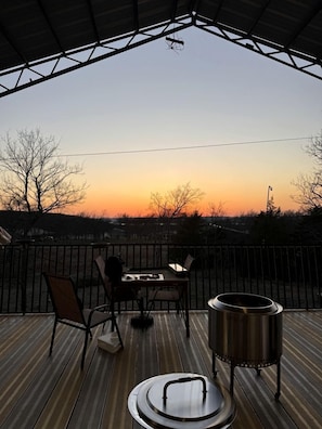 Eine Holzdielen-Terrasse mit Tisch, Stühlen, einem Grill und Blick auf den Sonnenuntergang.