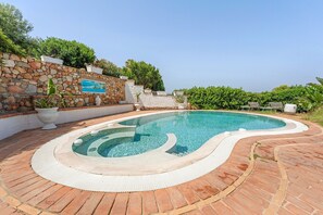 Piscina amb mur de pedra i plantes, envoltada per un paviment de teules.
