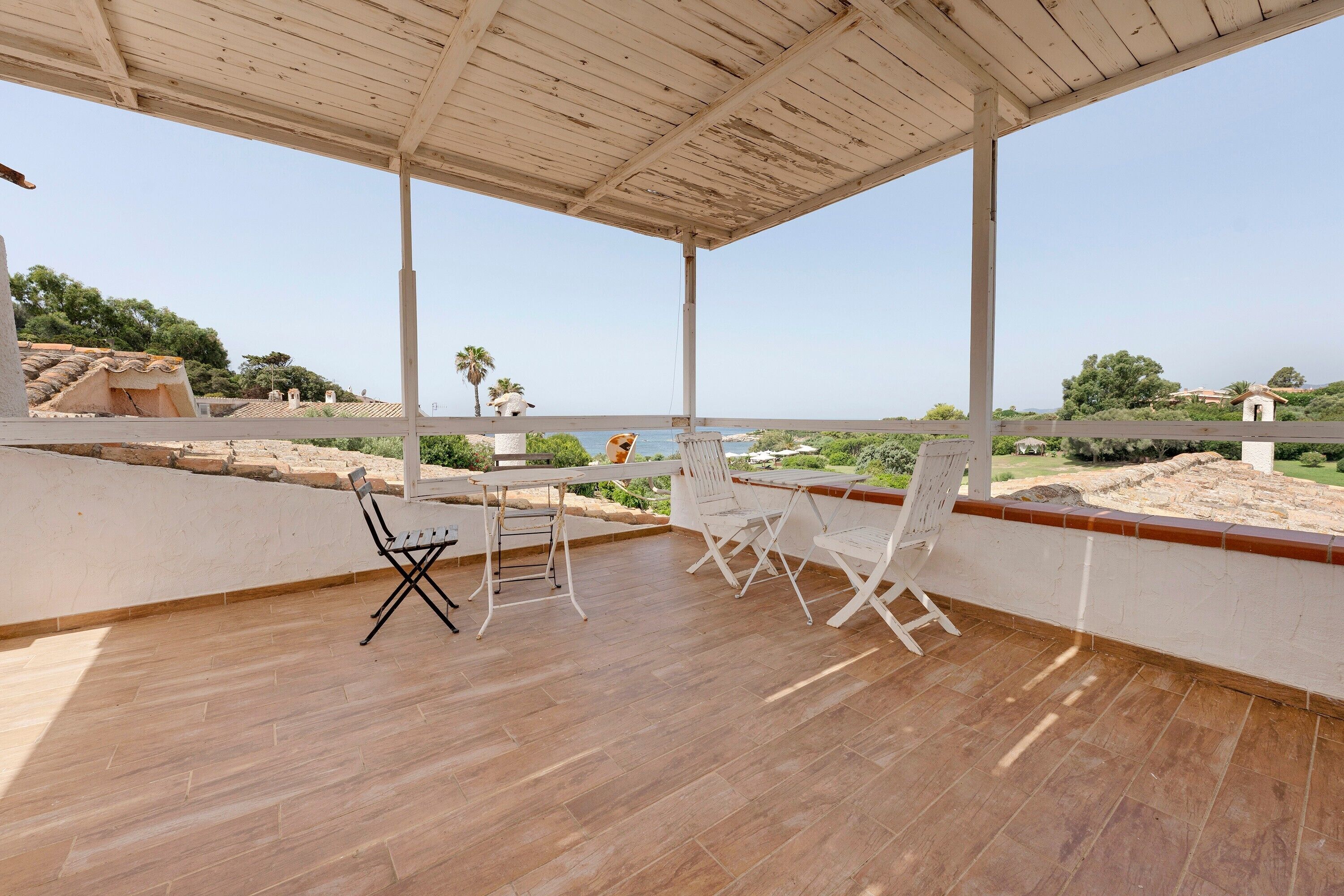 Un espacio al aire libre techado con suelo de madera, sillas de metal blanco y una mesa.