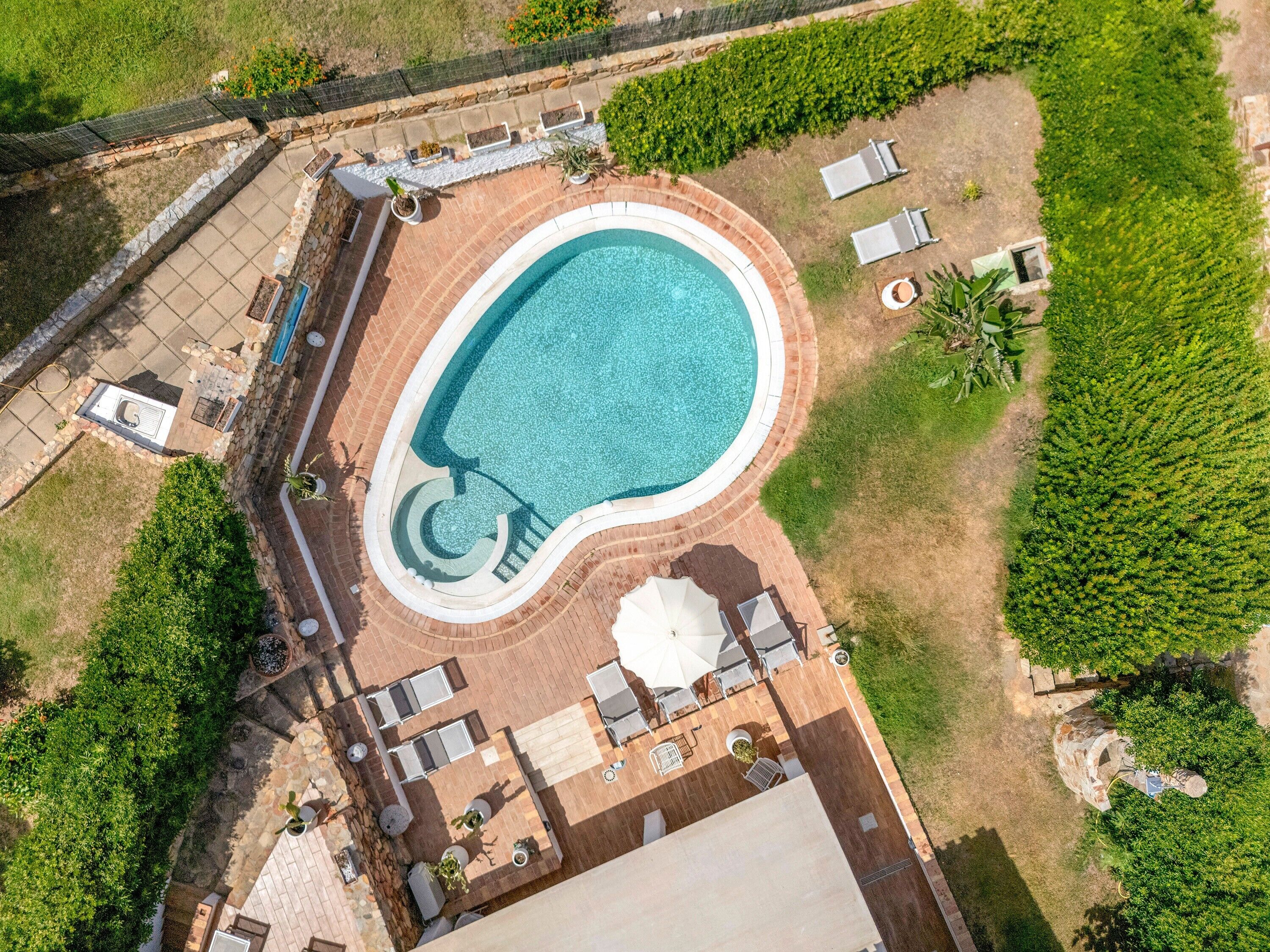 An aerial view of a property with a swimming pool, patio area with seating, and surrounding greenery.