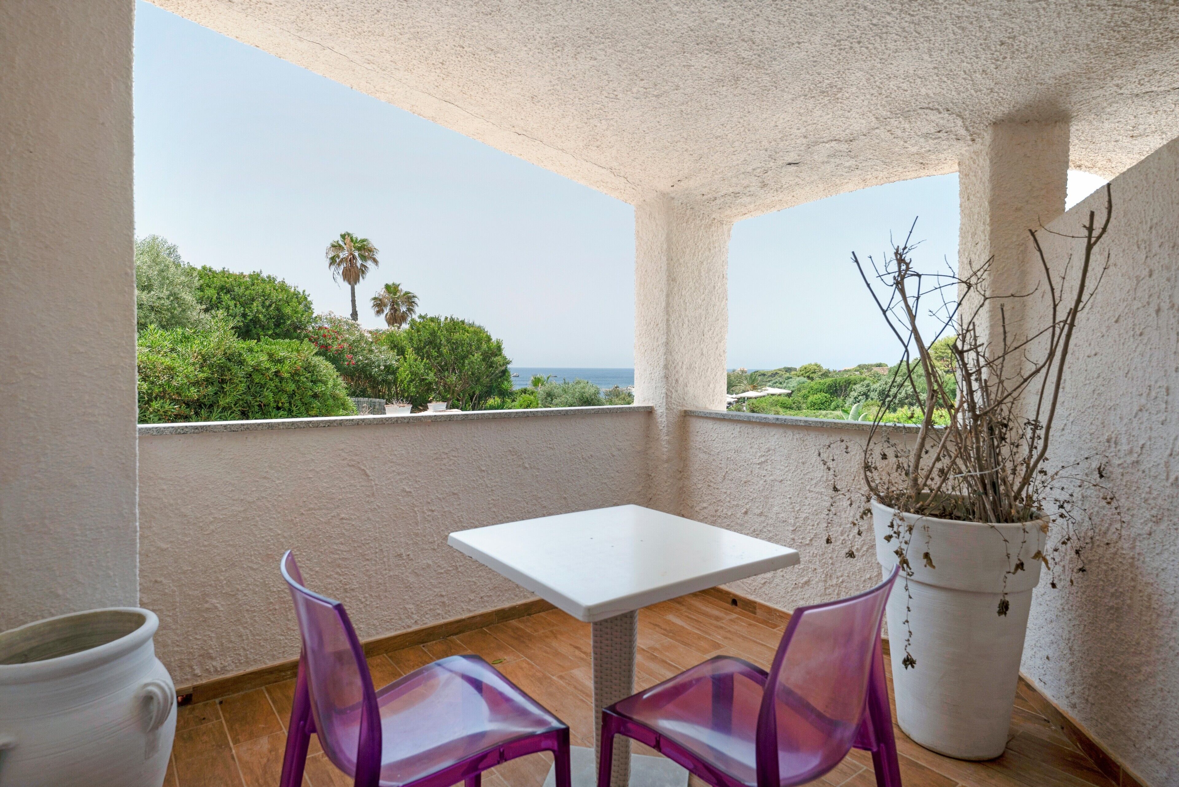 Un balcón con una mesa blanca y dos sillas moradas, con vistas a un paisaje costero con árboles y el mar.