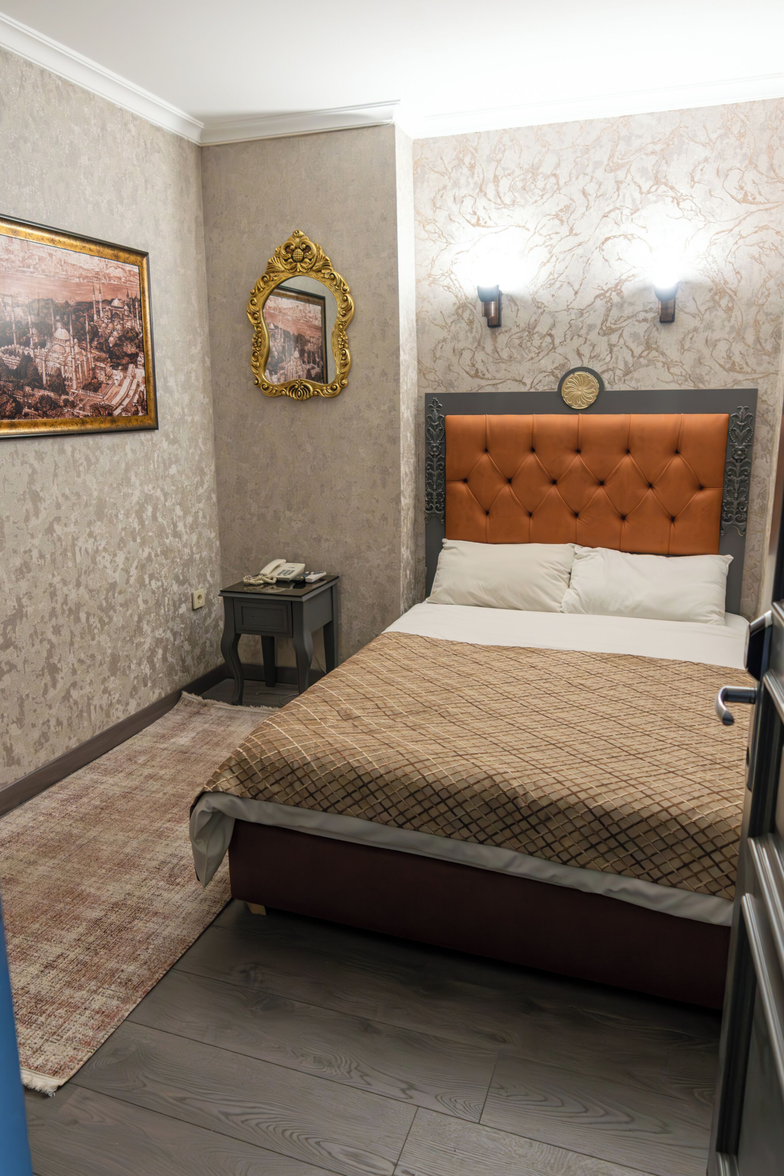 Een slaapkamer met een gestuukt hoofdeinde, een bed met een geborduurd sprei, een klein nachtkastje met een telefoon, en een lijst met een afbeelding aan de muur.