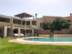 A pool area with lounge chairs and umbrellas, a stone wall, and a multi-level building with balconies.