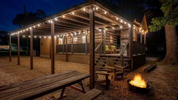 A wooden structure with a covered seating area, a fire pit, and string lights.