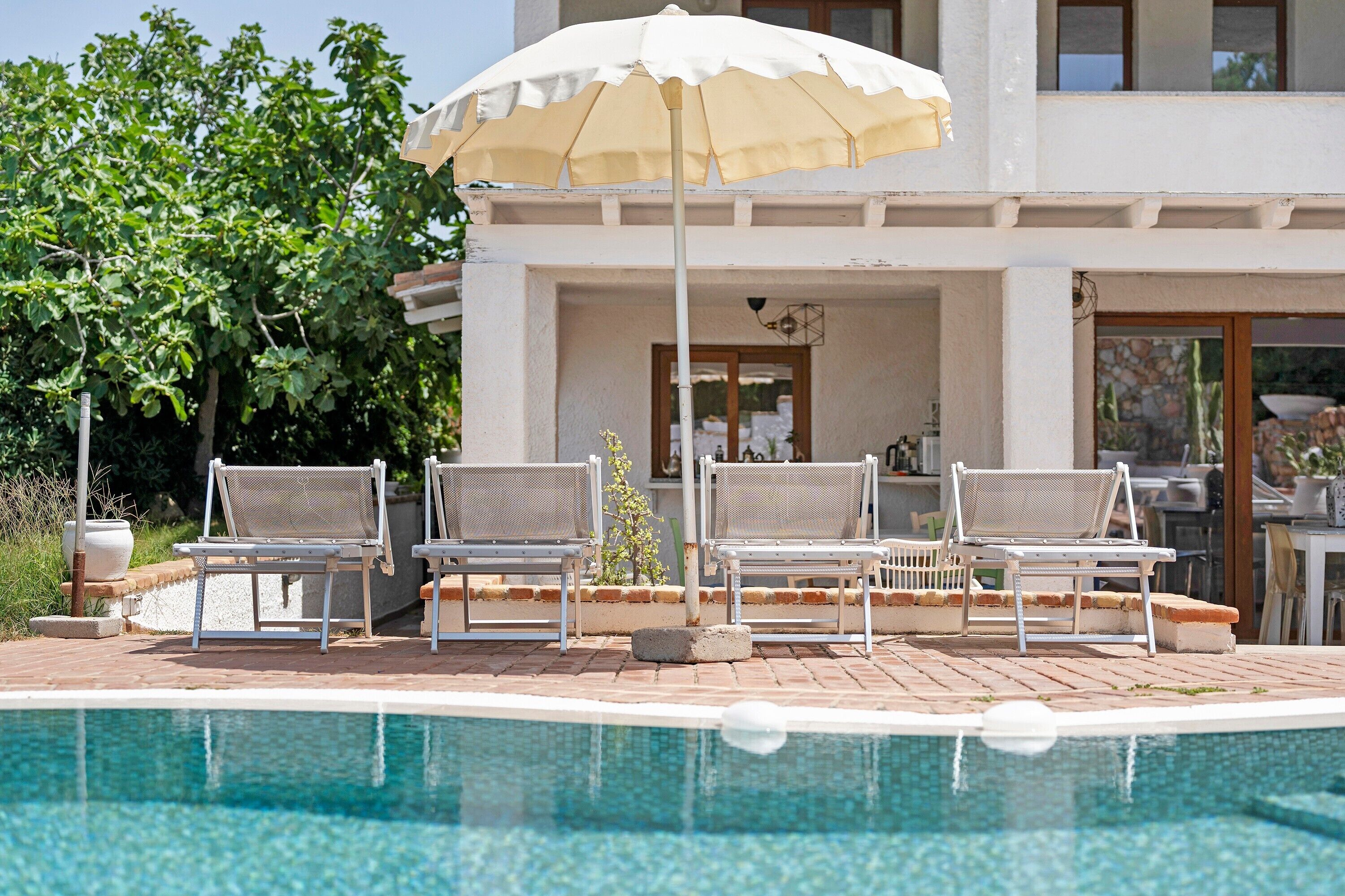 Zona piscina con poltrone da relax e un ombrellone bianco.