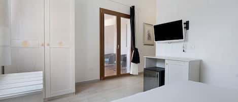 Habitación de hotel con cama, televisor montado en la pared, armario y puerta corredera de vidrio que da a un espacio exterior con mobiliario.