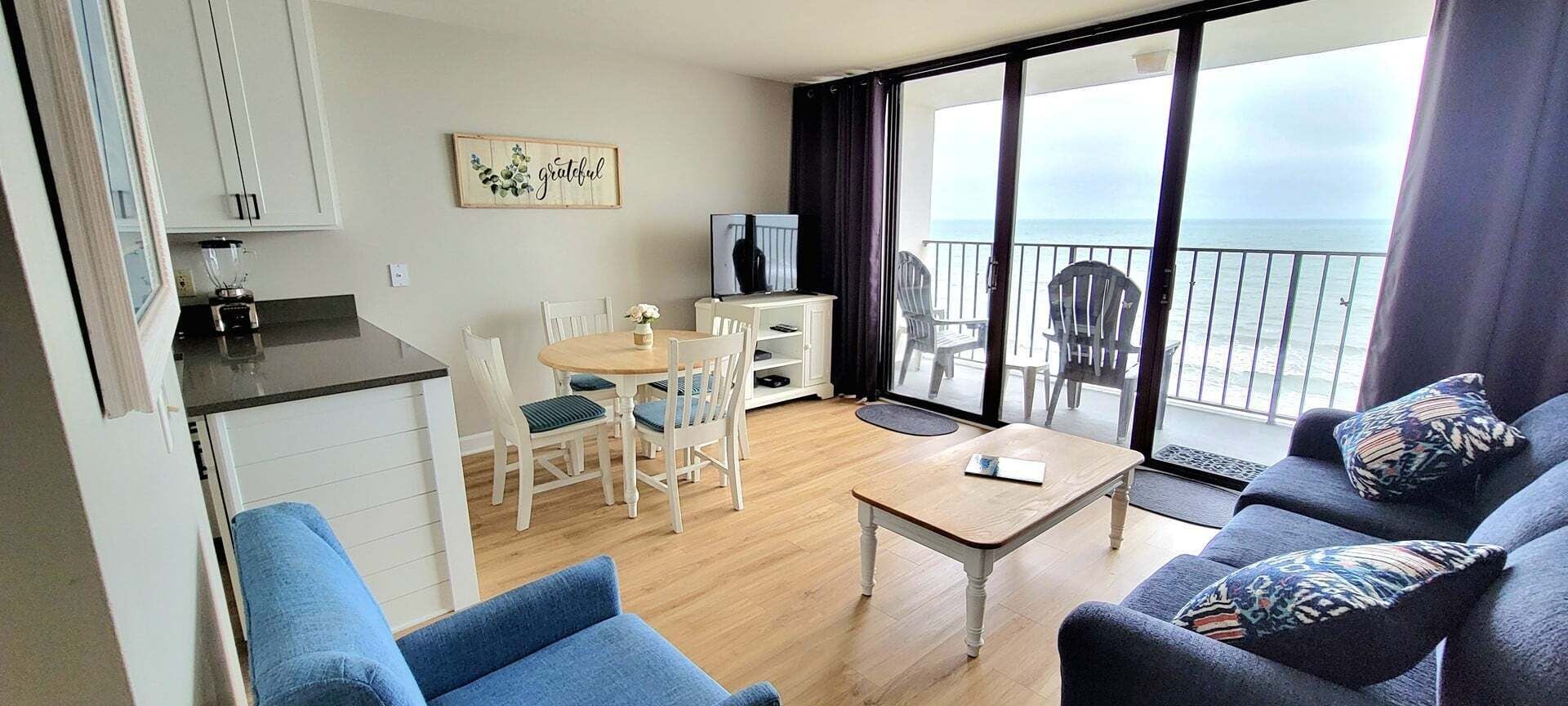 A room with a balcony, a dining area, a sofa, and a kitchen area.