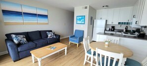 A living room with a sofa, a coffee table, and a dining table with chairs. There is a kitchen area with cabinets and appliances. - Oceanfront bliss awaits in this 1-bedroom condo 0804 (Carolina Beach)