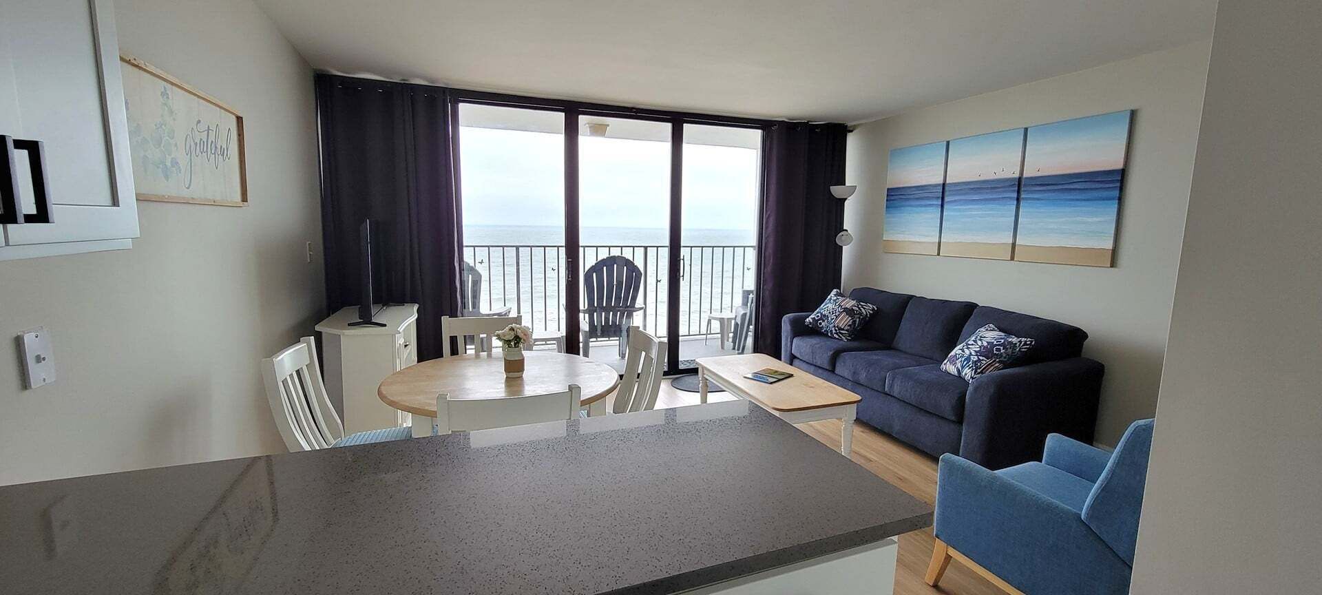 A living room with a sofa, a dining table with chairs, a balcony with a view of the sea, and a painting on the wall.