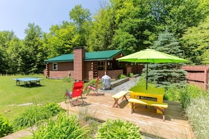 Une terrasse en bois avec une table de ping-pong, des chaises rouges, une table de pique-nique jaune et un parasol vert.