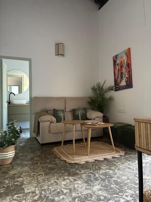 Ein Wohnzimmer mit einem beigen Sofa, einem kleinen Holztisch, einem gemusterten Teppich, einer Topfpflanze und einem Bild an der Wand.