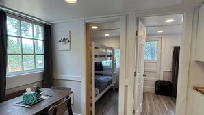 A room with a wooden floor, a dining table with chairs, a bunk bed, and a window with curtains.
