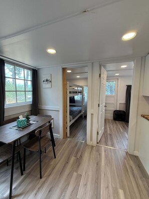 A room with a wooden floor, a dining table with chairs, a bunk bed, and a window with curtains.