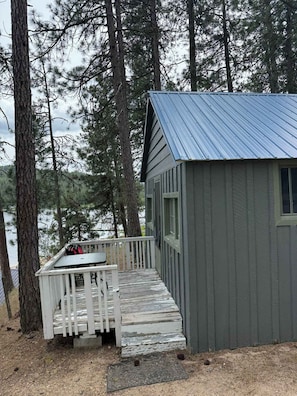 Una cabaña pequeña con terraza de madera y techo metálico, rodeada de altos pinos.
