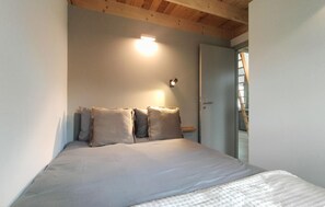 Une chambre à coucher avec un lit, des oreillers, une lampe fixée au mur et une porte donnant sur un escalier.