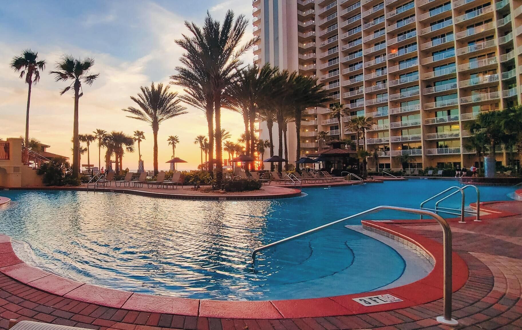 A swimming pool with lounge chairs and palm trees, surrounded by a high-rise hotel building.
