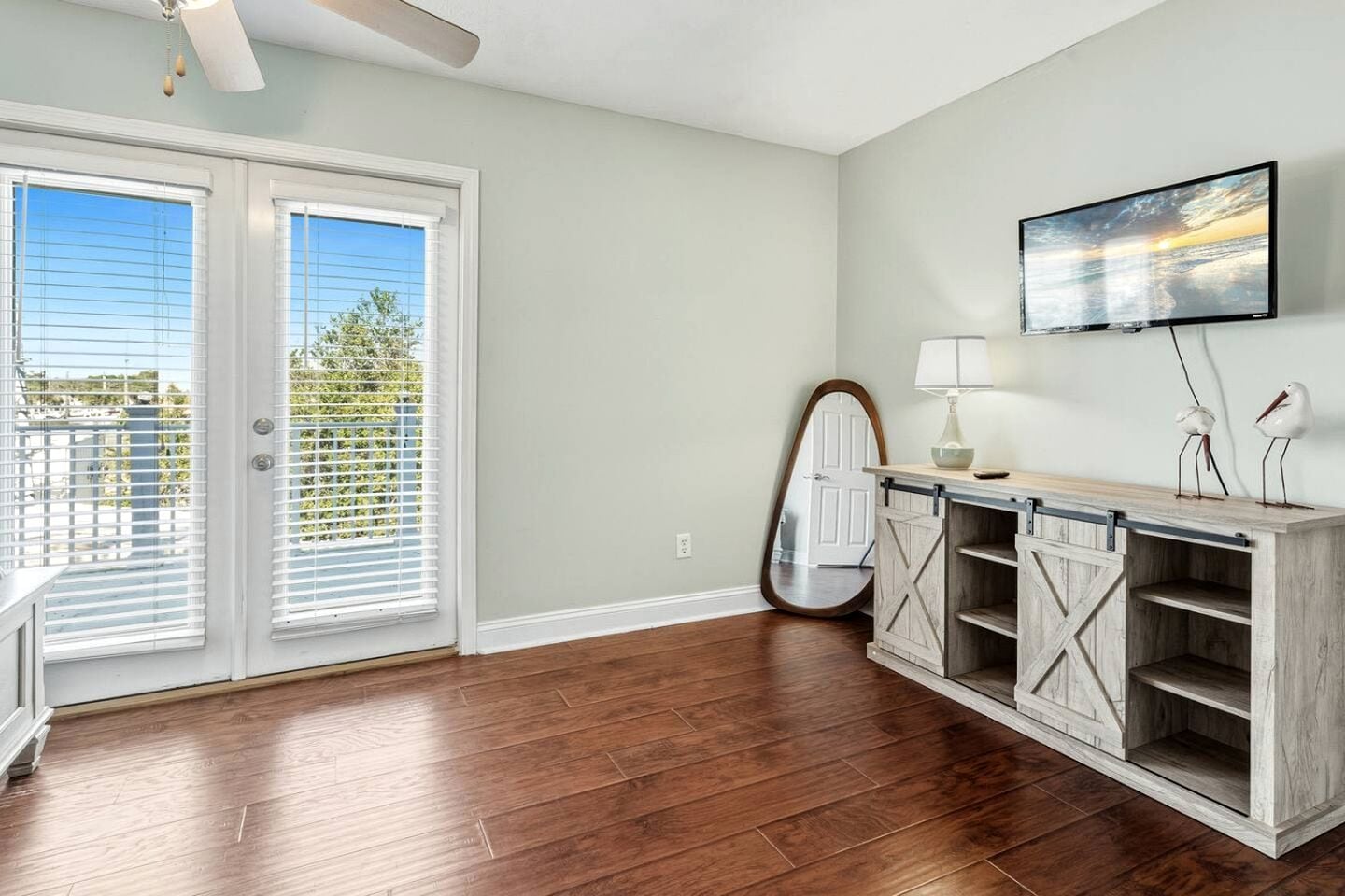 A room with a sliding glass door, a mounted TV, a wooden cabinet with shelves, and a ceiling fan.