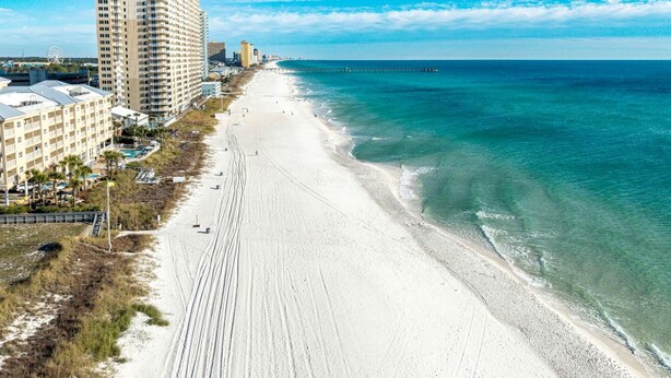 Uma praia com uma extensa faixa de areia, um píer que se projeta para o oceano e um hotel alto à esquerda.