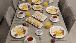Mesa de comedor servida con alimentos para el desayuno, incluyendo huevos, salchichas y tostadas, rodeada de sillas acolchadas.