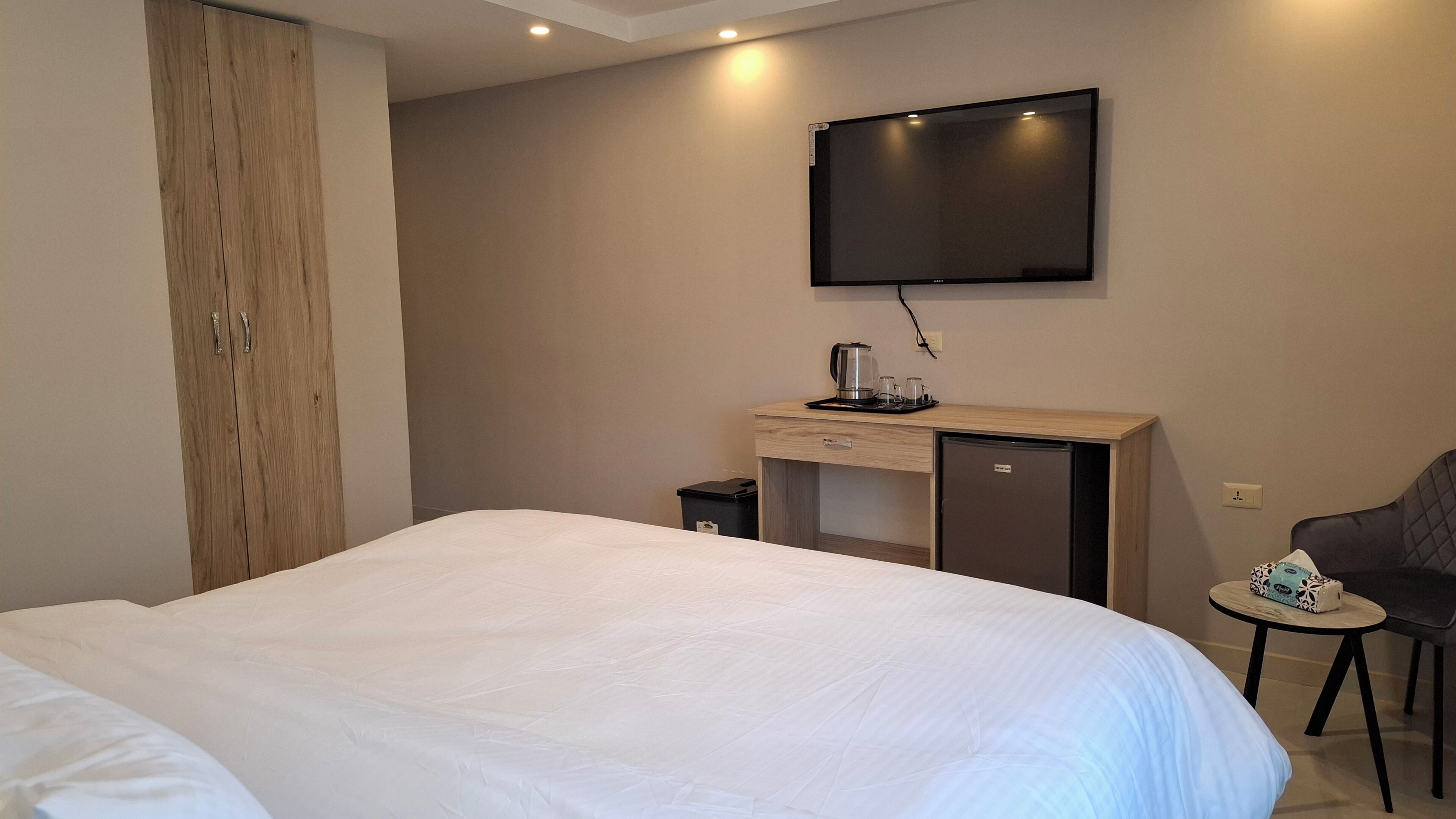 A hotel room with a bed, a desk with a television, a small refrigerator, a trash bin, and a chair.