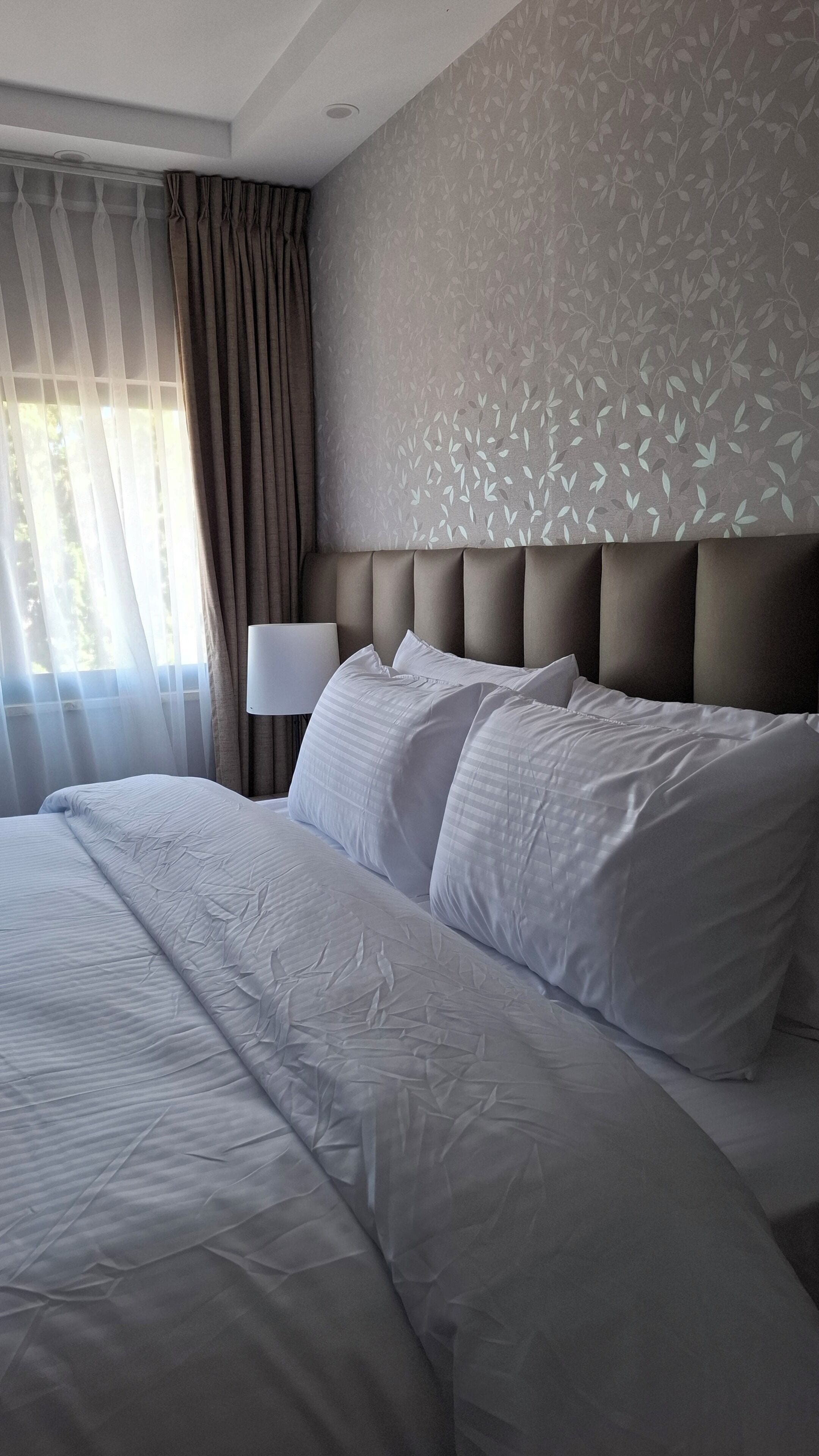 A hotel room with a large bed, a headboard, a nightstand with a lamp, and a window with curtains.