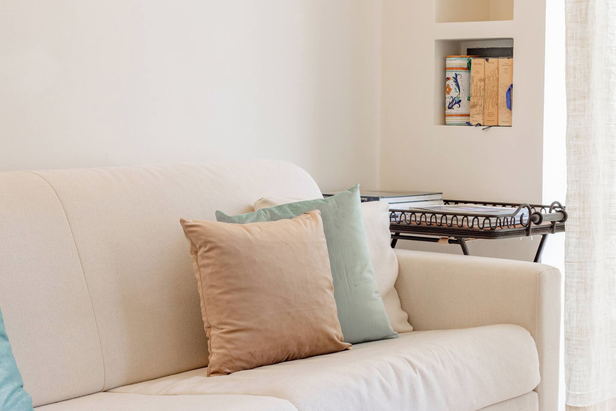 Una sala de estar con un sofá, almohadones, una mesita con libros, y una estantería con libros y adornos.