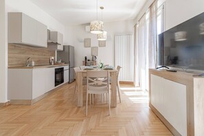 A modern kitchen with a dining area, wooden flooring, and a large TV on the right. - CASA MIMOSA - Parcheggio (Sanremo)