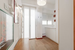 Un couloir avec un plancher en bois, des murs blancs, une porte dotée d’un miroir et un luminaire au plafond.