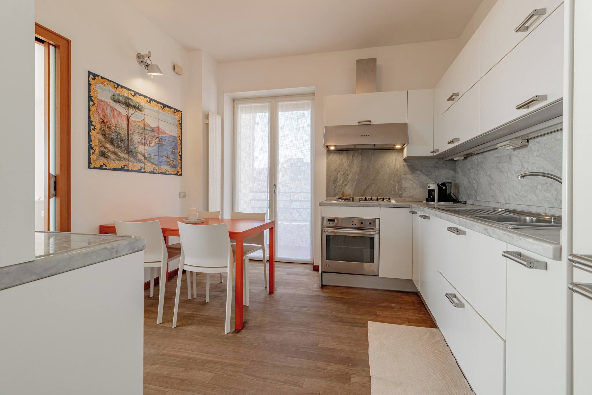 Una cocina moderna con armarios blancos, un comedor con mesa y sillas de madera, y un gran ventanal con cortinas ligeras.