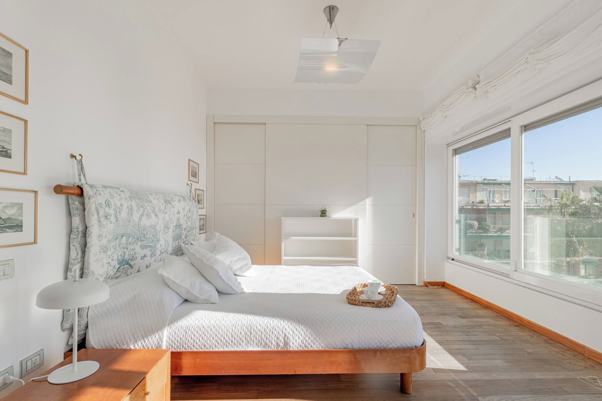 Une chambre moderne avec un lit en bois, du linge de lit blanc, une lampe de chevet, une vue sur la ville à travers de grandes fenêtres et une petite étagère avec une plante.