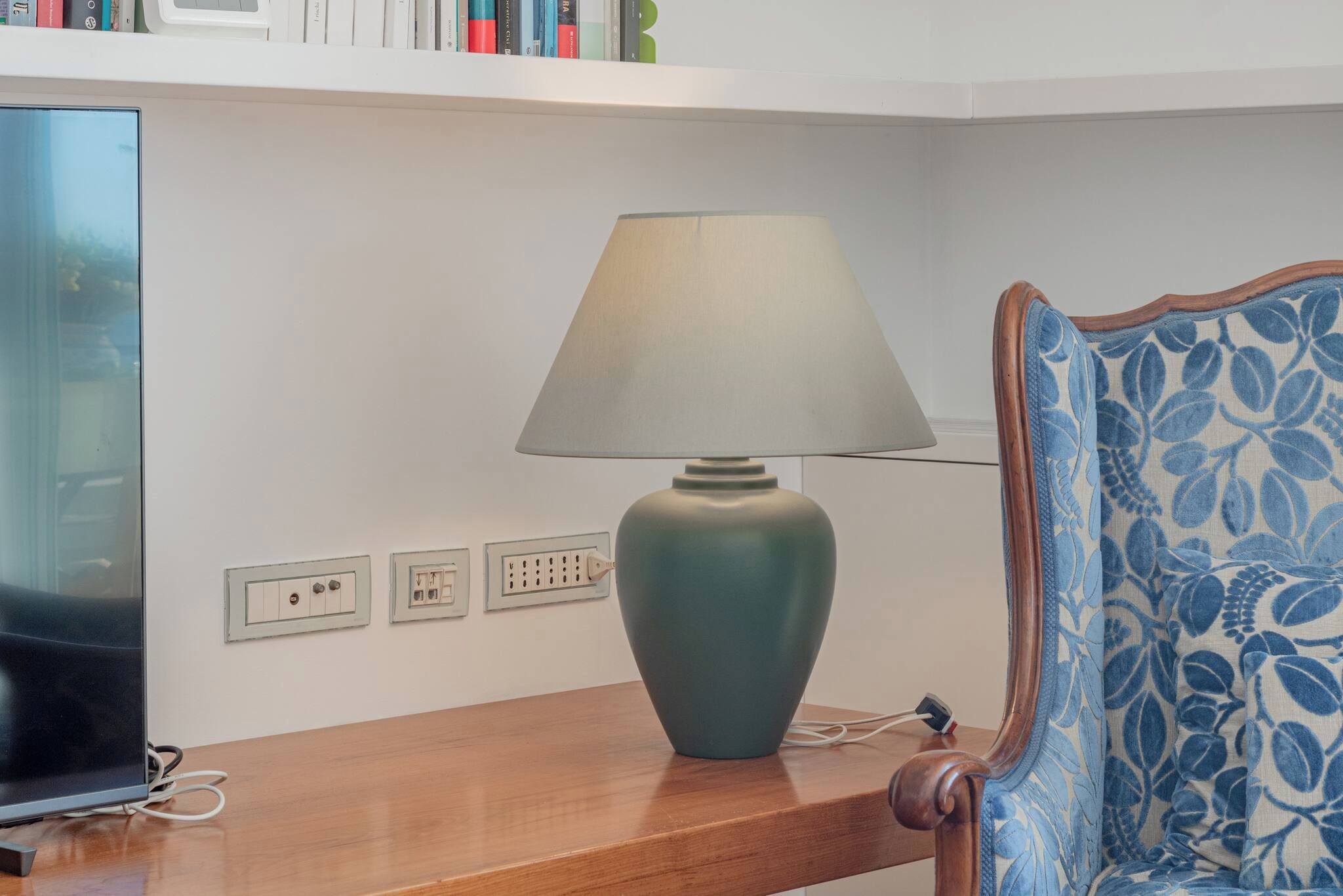 Una habitación con televisión, lámpara, una silla estampada con motivos florales y una estantería llena de libros.