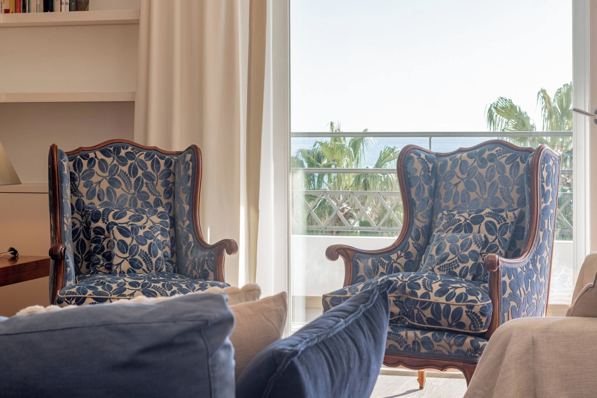 Une pièce comprenant deux fauteuils à motifs floraux, un canapé et une vue sur des palmiers depuis un balcon.